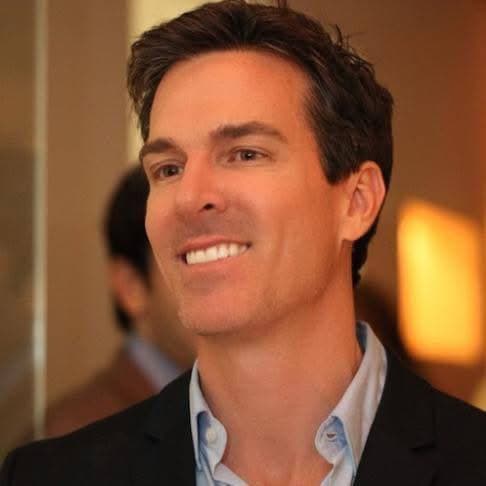 Smiling man with short dark hair wearing a black blazer over a light blue shirt.